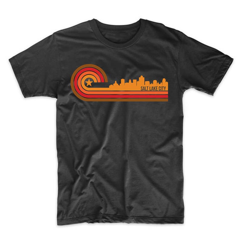 May include: A black t-shirt with a retro-style graphic of the Salt Lake City skyline in orange, red, and yellow. The graphic features a star and a curved line design.