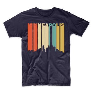 May include: A navy blue t-shirt featuring a vintage-style graphic of the Minneapolis skyline. The graphic has a multi-coloured striped background with the word "MINNEAPOLIS" in white letters.