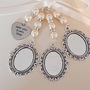 May include: A white satin ribbon tied in a bow, suspending three silver-toned oval photo frames and a silver heart-shaped charm engraved with the text With you Every Step. Each frame is attached to a strand of white faux pearls and rhinestones.