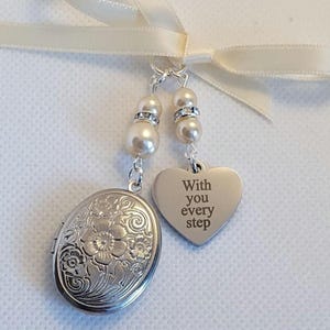 May include: A silver-toned oval locket with an embossed floral pattern and a heart-shaped charm engraved with the text With you every step. Both pieces are suspended from a cream-coloured ribbon by two silver chains adorned with white pearl beads.