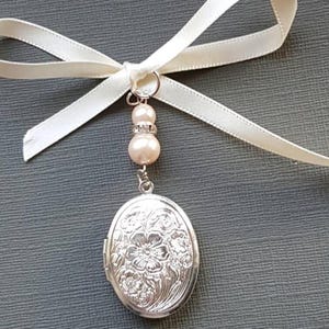 May include: An oval silver locket featuring an embossed floral pattern on the front. The locket hangs from a silver jump ring attached to two round cream-coloured beads and a small rhinestone spacer, finished with a cream satin ribbon bow.