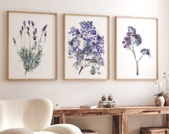 May include: Three framed watercolor paintings of purple flowers on a white wall above a wooden table with a white armchair and a small wooden table with a book on top.
