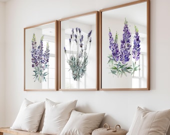 May include: Three framed watercolor paintings of purple lupine flowers with green stems and leaves. The paintings are displayed on a white wall above a wooden bench with cushions.