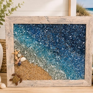 May include: A rectangular wall art piece in a weathered light wood frame featuring a beach scene created with tan sand and layers of crushed glass in shades of light blue, turquoise, and deep navy. A small brown turtle and starfish are attached.