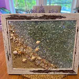 May include: A framed beach scene with a distressed white wooden frame. The artwork features sand, seashells, starfish, and small turtle figurines. The ocean is represented by green and blue crushed glass. Decorative sprigs are attached to the frame.