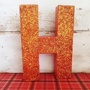 May include: Orange glitter-covered letter "H" on a red base. The letter is displayed against a white wooden background and a red plaid surface. The glitter gives the letter a sparkling appearance.