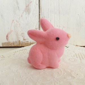 May include: A small, pink flocked bunny figurine with black eyes and a light pink nose. The bunny is sitting on a white lace doily, with a white wooden background.
