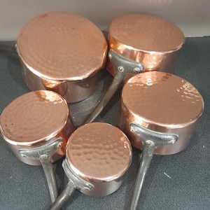 May include: Set of five copper pots with hammered surfaces and black handles. The pots are arranged in a pile, with the largest pot at the bottom and the smallest pot at the top.