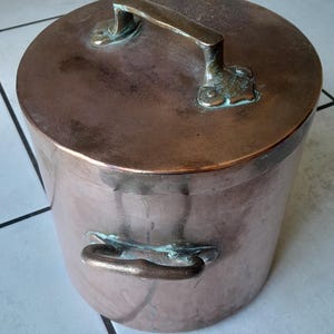 Può includere: Una pentola di rame con coperchio e due manici. La pentola ha una forma cilindrica e una superficie lucida. Il coperchio ha un manico grande e la pentola ha due manici laterali più piccoli. I manici presentano una patina verdastra.