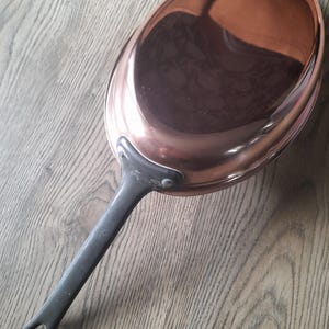 May include: A copper-colored oval frying pan with a long, dark gray handle. The pan's interior reflects the surrounding environment. The handle has a loop at the end for hanging. The pan is designed for cooking.