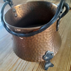 May include: A hammered copper pot with a black iron handle and three legs. The pot has a rustic, antique look.