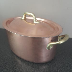 May include: An oval copper pot with a matching lid and two brass side handles. The pot features a polished metallic finish, a smooth rounded body, and a single brass handle centered on the lid. The construction shows visible rivets connecting the handles.