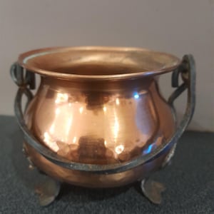 Peut inclure: Un petit chaudron en cuivre martelé de forme ronde avec une large ouverture et une finition métallique polie. Le récipient est soutenu par une structure en métal sombre et patiné, comprenant trois petits pieds et une anse incurvée.
