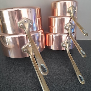 May include: A set of five polished copper saucepans of varying sizes, each featuring a shiny metallic finish and a long, curved brass handle securely attached with three visible rivets. The pans are stacked in two columns, showcasing their smooth, reflective surfaces and sturdy construction.