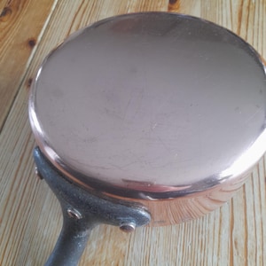 May include: A copper-colored saucepan with a dark metal handle. The pan has a rounded shape and a shiny surface. The handle is attached with rivets. The pan is sitting on a wooden surface.