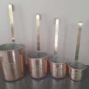 May include: Four copper ladles with brass handles hang against a grey wall. The ladles vary in size and have labels indicating their capacity: 1L, 050L, 025L, and OIL. The copper has a warm, reddish-brown hue.