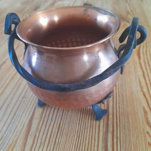 Può includere: Un piccolo calderone color rame con un corpo arrotondato e un'ampia apertura. Presenta due manici curvi in metallo nero e tre piccoli piedini decorativi. Il calderone ha una superficie lucida e riflettente.