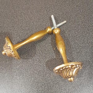 Op de afbeelding: Twee gouden metalen deurgrepen met sierlijke bloemmotieven. De grepen zijn bevestigd aan een metalen schroef.