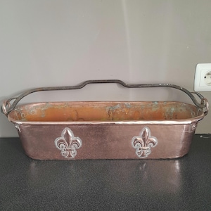 Puede incluir: Una maceta de cobre con dos diseños de flor de lis y un asa de metal negro.