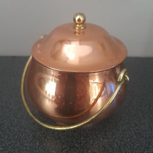 Puede incluir: Una pequeña olla redonda de cobre con acabado pulido, que presenta una tapa abovedada a juego rematada con un pequeño pomo de color latón. Un asa de metal fina y curvada de color latón está sujeta a los lados de la olla.