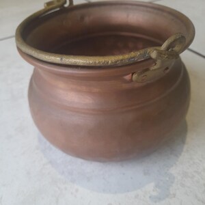 May include: A copper cauldron with a rounded base and a brass handle. The cauldron has a wide opening and a sturdy handle for carrying. The metal has a warm, reddish-brown hue, suggesting age and use. The cauldron is likely used for cooking or decorative purposes.
