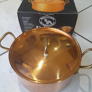 Puede incluir: Una cacerola Faitout de color cobre con tapa y asas de latón. La olla es redonda y tiene un acabado brillante. La caja tiene las palabras "Faitout" y "Les Cuivres de Fauconie" impresas.