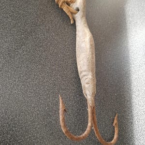 Puede incluir: Un gancho de pesca vintage con aspecto desgastado. El gancho presenta un mango claro en forma de pez con un agujero en la parte superior y dos ganchos curvos oxidados. Una cuerda anudada está unida al mango.