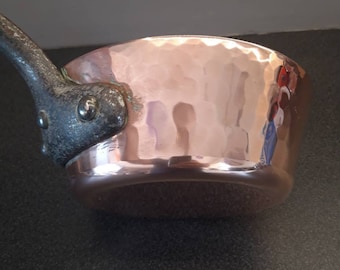 May include: A hammered copper saucepan with a black iron handle. The saucepan has a rounded bottom and a wide, shallow body.