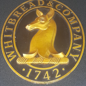 May include: A circular, black and gold sign with the text "Whitbread & Company" and the year "1742" in gold lettering. A gold stylized animal head is in the center of the sign.