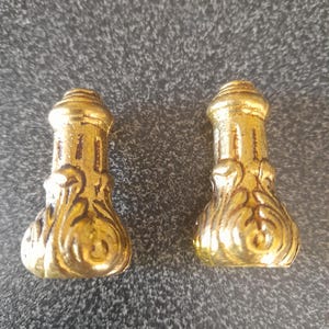 Puede incluir: Dos patas de mueble doradas y ornamentadas. Cada pata tiene una base bulbosa con adornos decorativos y una parte superior cilíndrica. Las patas parecen ser de metal y tienen una superficie brillante y reflectante.