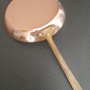 May include: A copper-colored frying pan with a long, brass-toned handle. The pan's interior is a light copper color, and the handle is attached with three rivets. The pan is set against a dark gray surface.