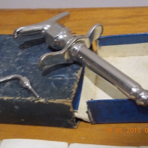 Puede incluir: Una jeringa dental plateada en una caja azul. La jeringa tiene un cañón largo y delgado y un mango con una palanca. La caja tiene una tapa azul y una etiqueta blanca con la fecha "29 05 2018 08:52".