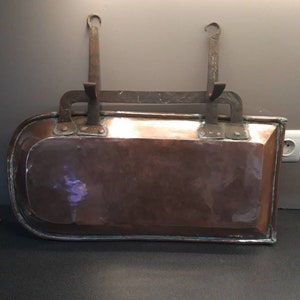 Può includere: Una teglia da forno in rame con una forma curva e due ganci di ferro per appenderla. La teglia ha una patina marrone scuro e una tinta verde intorno ai bordi.