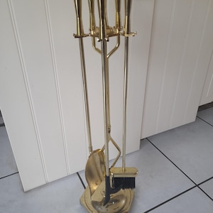 Könnte beinhalten: Ein Vintage-Kaminwerkzeug-Set aus Messing. Das Set enthält einen Schürhaken, eine Schaufel, eine Zange und eine Bürste, alle mit Messinggriffen und einem passenden Ständer. Die Werkzeuge sind auf einer sechseckigen Basis angeordnet.