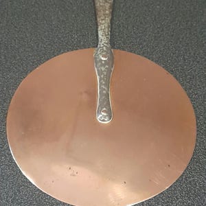May include: A round copper pan lid with a long, silver handle. The lid is slightly tarnished and has a few scratches.