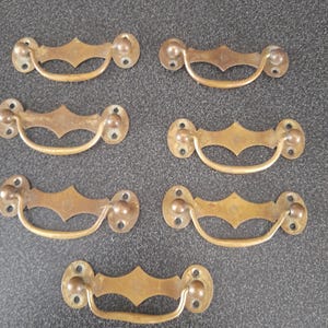 May include: Six antique brass drawer pulls. Each pull features a decorative diamond-shaped center and a curved handle. The pulls have a warm, aged patina, suggesting vintage or reclaimed hardware. The mounting plates have two screw holes each.