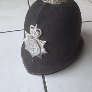 May include: A black felt custodian helmet featuring a silver-toned metal badge on the front with a crown emblem and the text Avon and Somerset Constabulary. The helmet includes a silver-toned metal top rose ornament and two side ventilation eyelets.