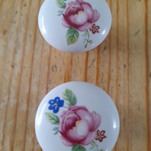 May include: Two white ceramic cabinet knobs with a floral design. Each knob features a large pink rose, blue and red accent flowers, and green leaves. The knobs are round and have a glossy finish.
