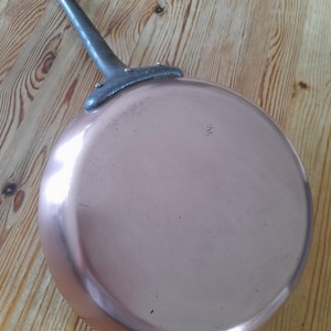Puede incluir: Una sartén de color cobre con un mango largo y gris. La sartén es redonda con un borde ligeramente curvado. El mango está sujeto a la sartén con remaches. La sartén está sobre una superficie de madera.