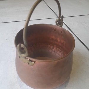 Puede incluir: Una pequeña olla de color cobre con un asa y detalles de latón. La olla tiene un cuerpo redondeado y una textura martillada en el interior. El asa es arqueada y se fija a la olla con accesorios de latón. Un artículo de cocina vintage.