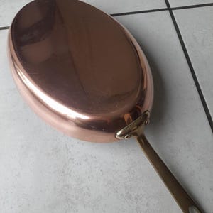 Könnte beinhalten: Eine ovale Bratpfanne aus Kupfer mit einem langen Messinggriff. Die Pfanne hat eine polierte, reflektierende Oberfläche und ist zum Kochen bestimmt. Der Griff ist mit Nieten befestigt und hat eine Schlaufe zum Aufhängen.