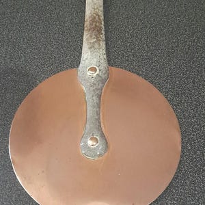 May include: A vintage copper pan lid with a long, silver handle. The handle has a loop at the top and two decorative rivets.