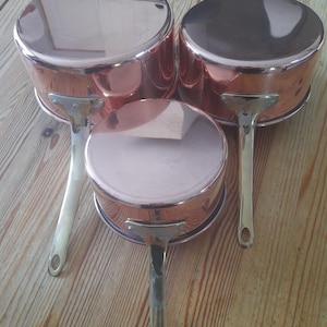 May include: Three copper saucepans with silver handles are arranged on a wooden surface. The pans vary in size, with a reflective surface. The copper has a warm, reddish-brown hue, and the handles are attached with rivets.