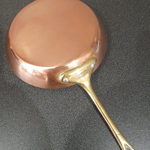 Puede incluir: Sartén de color cobre con un mango de latón. La sartén tiene forma redonda y una superficie brillante. El mango está remachado y tiene un lazo para colgar. Utensilios de cocina.