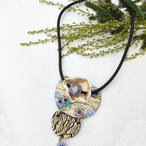 K&ouml;nnte beinhalten: Eine goldene und blaue Halskette mit einem mehrschichtigen Anh&auml;nger, der eine wei&szlig;e Blume, blaue und goldene Kreise und eine blaue Perle zeigt. Die Halskette besteht aus schwarzem Band.