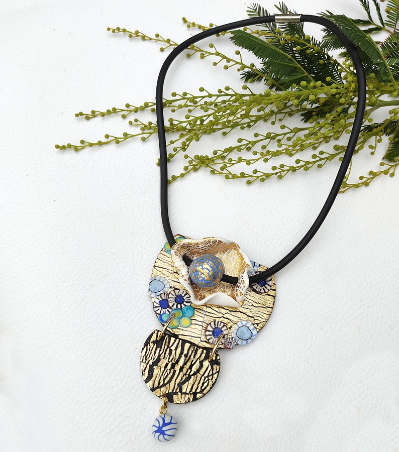 K&ouml;nnte beinhalten: Eine goldene und blaue Halskette mit einem mehrschichtigen Anh&auml;nger, der eine wei&szlig;e Blume, blaue und goldene Kreise und eine blaue Perle zeigt. Die Halskette besteht aus schwarzem Band.