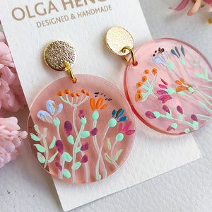 Peut inclure: Une paire de boucles d'oreilles rondes en acrylique rose avec des clous dorés. Chaque boucle d'oreille présente un motif floral peint à la main dans des tons de bleu, orange, violet et vert. Les boucles d'oreilles sont conçues et fabriquées à la main.