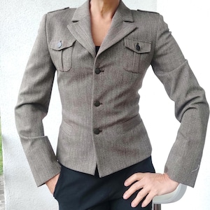 Peut inclure: Un blazer taupe avec un motif à chevrons subtil, doté d'un revers cranté, d'épaulettes et de deux poches poitrine à rabat boutonné. La veste se ferme avec trois boutons foncés sur le devant et comprend deux poches passepoilées inférieures.