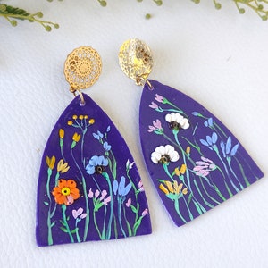 May include: Purple polymer clay earrings with a floral design. The earrings feature a variety of flowers in shades of blue, white, yellow, orange, and pink. The earrings are attached to gold-toned filigree earring hooks.