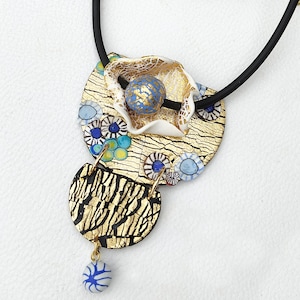 K&ouml;nnte beinhalten: Eine goldene und schwarze Halskette mit einem Anh&auml;nger in Form einer wei&szlig;en Blume mit blauen und goldenen Akzenten. Der Anh&auml;nger ist aus Polymer Clay gefertigt und hat eine strukturierte Oberfl&auml;che.